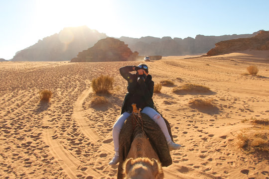 A woman rides a camel near dried bushes through the Wadi Rum desert in Jordan and simultaneously tries to take pictures. Sandstone mountains in the background.