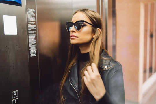 Portrait of sexy young caucasian woman fashion model girl standing by the modern elevator wearing black coat holding hair looking away eyeglasses waiting