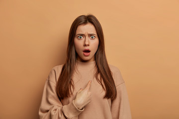 Stunned brunette European woman indicates at herself with indignant stupefied expression, shocked...