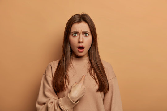 Stunned brunette European woman indicates at herself with indignant stupefied expression, shocked being chosen, frowns eyebrows, wears loose sweatshirt, models over beige wall. You blame me?