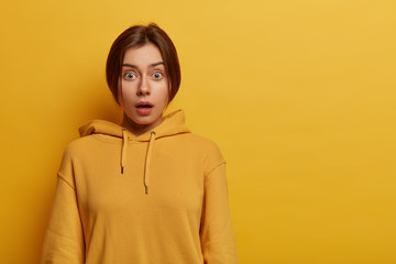 Portrait of astonished beautiful female hipster holds breath from excitement and surprisement, stares with bugged eyes, wears yellow casual hoodie, stands indoor alone, concerned by terrible news