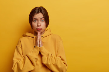 Dissatisfied woman has faith in better, holds hands in pray, asks for favor, has sad expression, supplicates and anticipates for something, dressed in sweatshirt, isolated over yellow background