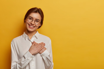 Charming positive European woman presses hands to chest, expresses gratitude for gift, appreciates help, wears round glasses and white shirt, stands against yellow background, looks with admiration