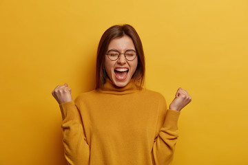 Fototapeta Overjoyed brunette young lady clenches two fists, celebrates success and exclaims with joy, shouts good job, makes hooray gesture, triumphs from successful deal or awesome news, feels thrilled obraz
