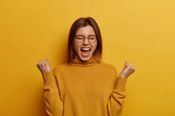 Fotobehang Overjoyed brunette young lady clenches two fists, celebrates success and exclaims with joy, shouts good job, makes hooray gesture, triumphs from successful deal or awesome news, feels thrilled © Wayhome Studio