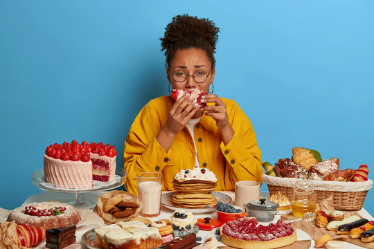 Gluttony and overeating concept. Upset crying ethnic woman eats piece of cake reluctantly, sits at table with many desserts, isolated over blue wall, feels hungry and greedy, wears yellow jacket