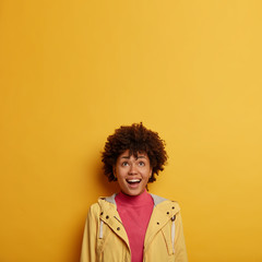 Obraz na płótnie Canvas Happy laughing woman looks above with interest and joy, checks out funny advertisement, dressed in casual jacket, has curly hair, poses against yellow background, feels amused, concentrated upwards