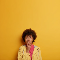 Fototapeta na wymiar Pensive dreamy woman with Afro hair concentrated above, makes decision, looks up thoughtfully, keeps hand near mouth, wears yellow anorak, poses in studio, copy space for your advertising or text