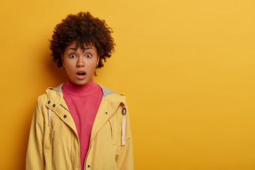 Obraz na płótnie Canvas Concerned frightened curly adult woman feels fear and panic because of terrible accident, keeps mouth opened, dressed in casual anorak, gasps amazed, isolated on yellow wall, copy space area