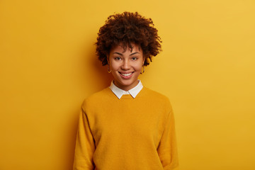Fototapeta na wymiar Portrait of beautiful tender woman has cheerful smile, wears casual jumper with white collar, stands against vivid yellow background, looks directly at camera, expresses good emotions, enjoys talk