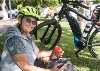 Naklejka premium Cheerful cyclist woman with the yellow helmet has a break in the park. Sitting in the grass and eating a red apple. Relax for an elderly person who watches the mobile phone