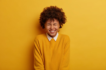 Obraz premium Photo of joyful emotional dark skinned woman squints face, smiles broadly, feels amused and entertained during party, wears casual jumper, models over bright yellow background. Emotions concept