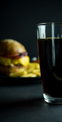 Close up of glass of cola near blurred burger and french fries on black background 