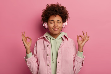 Relaxed beautiful woman makes zen gesture, meditates while listens peaceful music in headphones,...