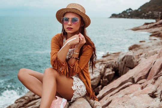 Lady dressed in boho style, straw hat, cape, bracelets and pendant made of eco wood, glasses, flash tattoos. Girl stands on a stone coast against the background of the sea, sensual portrait
