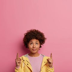 Vertical image of curious woman with Afro hair points above with both index fingers, demonstrates copy space upwards, wears windbreaker, poses and gestures against pink background. Promotion