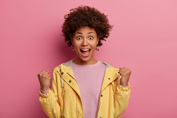 Obraz na płótnie Canvas Attractive emotional woman excited and glad to achieve victory, exclaims and clenches fists, being successful person, supports someone, stares at camera, isolated on pink background, roots for team