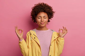 Patient relaxed dark skinned woman meditates and stands in yoga pose, keeps eyes closed, relaxes...
