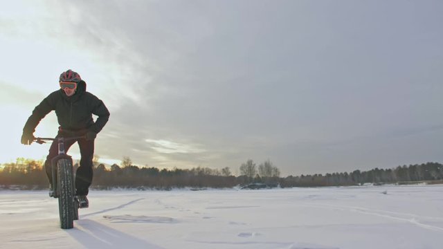 Professional extreme sportsman biker riding fat bike in outdoors. Cyclist ride in winter on snow ice. Man does trick on mountain bicycle with big tire in helmet and glasses. Slow motion in 180fps.
