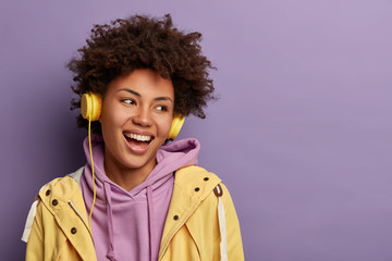 Close up shot of beautiful woman enjoys spare time with favourite music, gets ready for walk in park, smiles with teeth and looks aside, dressed in casual sweatshirt, isolated on purple background