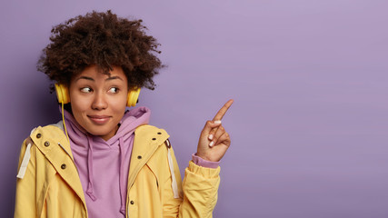 Pretty curly woman wears modern headphones listens music and advertises something, points fore...