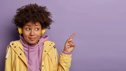 Schilderijen op glas Pretty curly woman wears modern headphones listens music and advertises something, points fore finger at upper right corner, wears casual outfit, isolated on violet background, uses stereo accessory © Wayhome Studio
