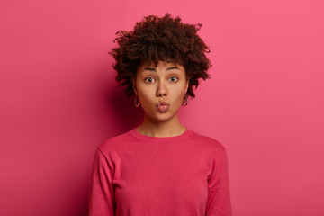 Lovely romantic dark skinned woman folds lips and awaits for kiss, has flirty expression, wears pink sweater, stands indoor, sends mwah at camera, isolated on pink background. Face expressions