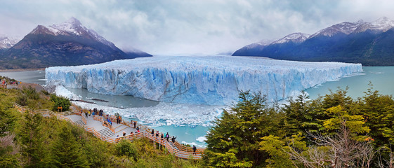 Panoramic view of Perito Moreno Glacier surrounded by green vegetation and snowy mountains, in El Calafate, Argentina, against a grey and cloudy sky.