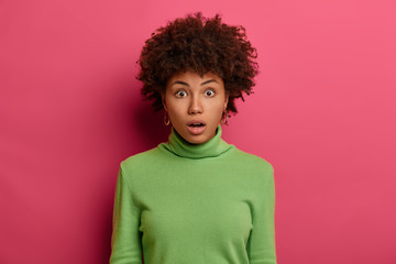 Surprised curly haired woman checks out something awesome and shocking, hears amazing news, wears green neck sweater, realizes terrible relevation or latest rumors, poses against pink studio wall.