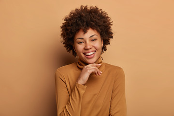 Obraz na płótnie Canvas Portrait of cute African American woman gazes with tenderness and delight, keeps hands under chin, laughs at positive funny story wears casual brown turtleneck models indoor recalls something pleasant
