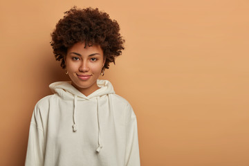 Obraz na płótnie Canvas Studio shot of good looking woman has Afro hair, has direct gaze and tender smile, wears white sweatshirt, poses over beige wall with copy space for your promotion. Human facial expressions.