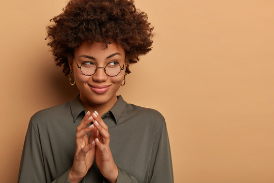 Portrait of attractive curly haired woman has tricky plan in mind, steeps fingers and looks mysteriously aside, wears round glasses and shirt, thinks about good idea for project, cunny look.