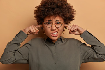 Human reactions concept. Headshot of Afro American woman complains on loud music, smirks face with displeasure, plugs ears, wears round spectacles and shirt, ignores noise, isolated on brown wall