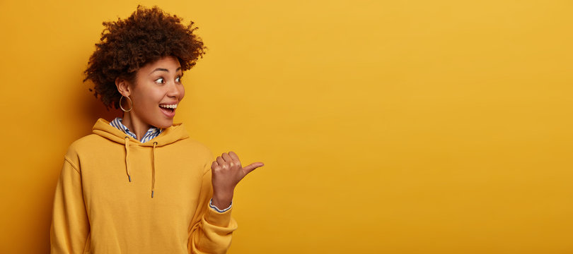 Optimistic glad Afro American hipster girl points thumb aside on blank space, shows awesome advertisement, expresses happiness, wears casual hoodie, isolated over yellow background, enjoys spare time