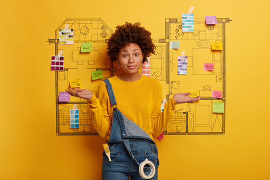 Hesitant woman architect poses against drawing sketch, spreads palms sideways, makes decision how to remodel house better, wears casual yellow sweater, denim overalls with paint brush in pocket - Powered by Adobe
