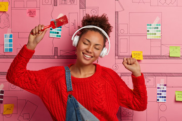 Amused relaxed woman dances happily, raises arms with paint brush, listens music in wireless...