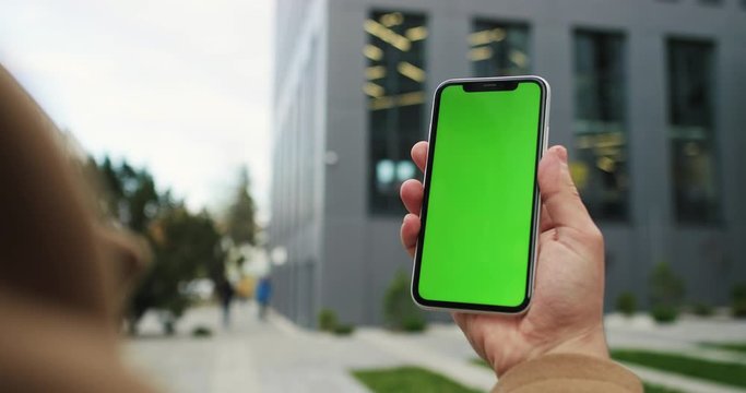 Male Caucasian hand rising up his black smartphone with green screen vertically while being outside at the building on the street. Chroma key.