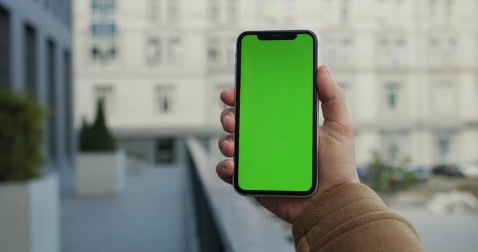 Vertical black phone with chroma key on the screen is in the female Caucasian hand outside at the city street. Green screen. Close up.