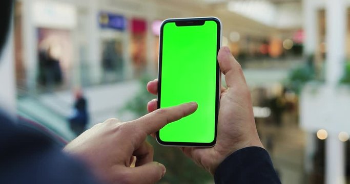 Close up of the black vertical smartphone with green screen in Caucasian male hand while he scrolling pictures on it in the big shopping mall. Indoors. Chroma key.
