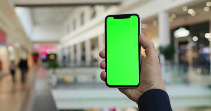 Close Up Of The Hand Of Caucasian Man Which Holding Vertical Black Phone With Green Screen Like He Doing Photo Of The City Shopping Mall Passage. Chroma Key. Indoor.