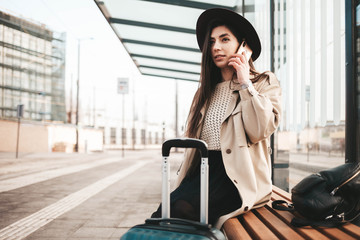 Fototapeta premium Young beautiful traveler girl is waiting at the public transport stop talking on the smart phone