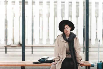 Young traveler lady in coat and hat with a sad face sits at a bus city stop alone