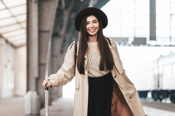 Portrait of young beautiful woman traveler in casual coat and hat who is waiting for a train on the platform of the railway station