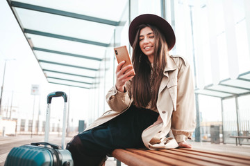 Beautiful positive girl traveler in coat and hat on his head sitting at a bus stop waiting for a...
