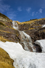 Fototapeta na wymiar Large, scenic, beautiful waterfall water falling from red rocky cliff under the snow during transition period between winter and spring