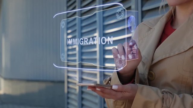 Unrecognizable businesswoman interacts HUD hologram with text Immigration. Woman in the coat uses the technology of the future mobile screen on the background of the city