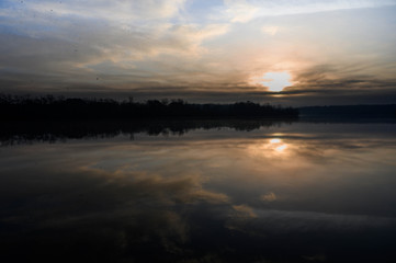 The glow of the morning sun has risen above the clouds. The reflections of the clouds and trees are shown on the calm still waters.