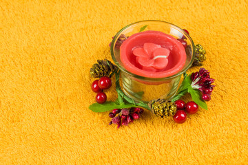 Red candle in the shape of a flower in a glass candlestick and decorated with cones on a yellow towel.