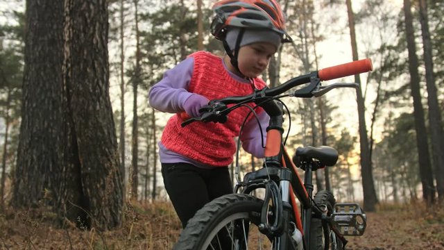 Kid wear helmet. One caucasian children rides bike road in autumn park. Little girl riding black orange cycle in forest. Biker motion ride with backpack and helmet. Mountain bike hardtail.