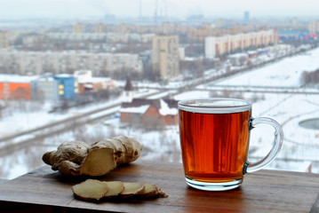 A transparent glass mug of hot black tea and sliced ​​ginger root close-up stand on a wooden surface against a blurry background of the winter panorama of the city of Togliatti.
